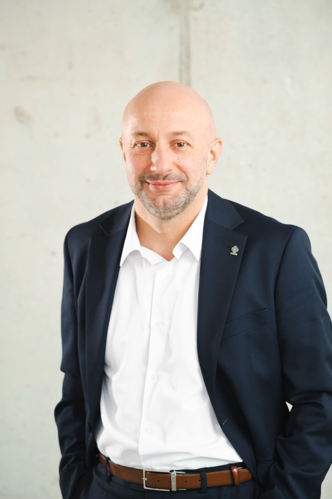 Hubert Gęsiarz w granatowej marynarce i białej koszuli, portret na tle betonowej ściany